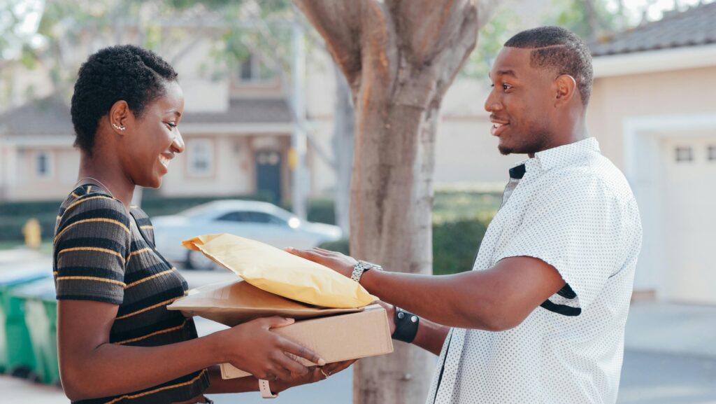A courier delivers packages to a smiling woman at an outdoor location, representing online shopping and delivery services.